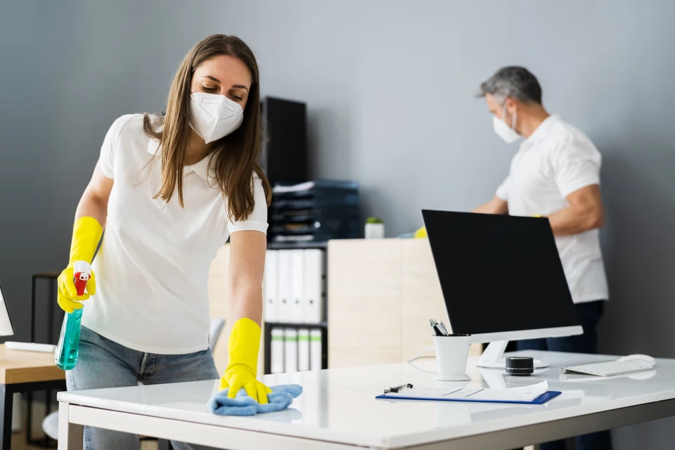 Femme en tenue de nettoyage professionnelle désinfecte un bureau moderne avec spray et chiffon, portant masque FFP2 et gants jaunes