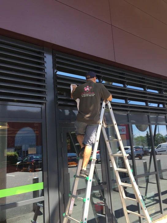 Ouvrier en combinaison bleue et harnais de sécurité rouge travaillant sur nacelle élévatrice jaune devant immeuble de bureaux moderne avec façade vitrée